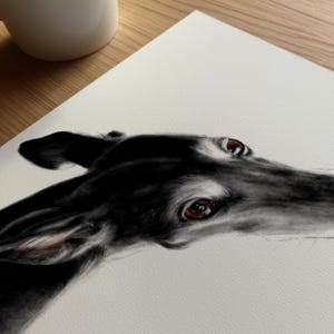 In the image: A detailed, black-and-white drawing of a greyhound's head. The dog's smooth, black coat contrasts with its brown eyes and the white paper. The artwork captures the dog's focused gaze and elegant features.