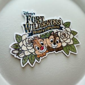May include: A sticker featuring Chip and Dale from Disney's Fort Wilderness Resort & Campground. The sticker has a white background with the text "Disney's Fort Wilderness Resort & Campground" in a banner. Chip and Dale are smiling, surrounded by white flowers and green leaves.