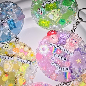 Puede incluir: Una colección de llaveros de resina coloridos con varios abalorios y cuentas. Cada llavero presenta un diseño único, incluyendo caras sonrientes, patos y las palabras "MAGIC MAKER" y "HAPPILY EVER AFTER".