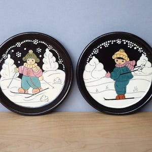 May include: Two round black decorative plates featuring hand-painted winter scenes. Each plate depicts a child wearing a hat, scarf, and winter clothing while skiing on a white snowy landscape with stylized white trees and falling snowflakes.