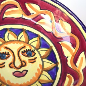 May include: A yellow ceramic plate featuring a hand-painted design of a sun with a smiling face, blue eyes, and a red mouth. The sun is surrounded by a blue and yellow floral pattern on a red background.