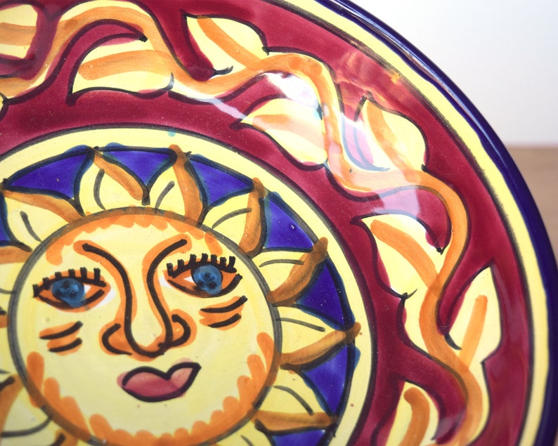 May include: A yellow ceramic plate featuring a hand-painted design of a sun with a smiling face, blue eyes, and a red mouth. The sun is surrounded by a blue and yellow floral pattern on a red background.