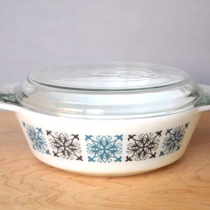 Puede incluir: Una fuente para horno blanca vintage con tapa de cristal transparente. El plato presenta un patrón repetido de diseños florales azules y negros. Tiene dos asas blancas.