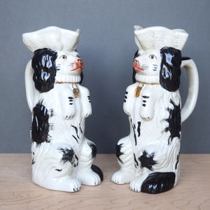 Puede incluir: Dos jarras de cerámica en forma de perro blanco y negro con un sombrero blanco en la parte superior. Las jarras tienen detalles dorados y están sobre una superficie de madera clara.