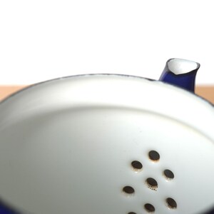 K&ouml;nnte beinhalten: Eine blaue Emailleteekanne mit wei&szlig;em Innenraum und einem Sieb mit mehreren L&ouml;chern. Die Teekanne ist aus Metall und hat ein klassisches Design.