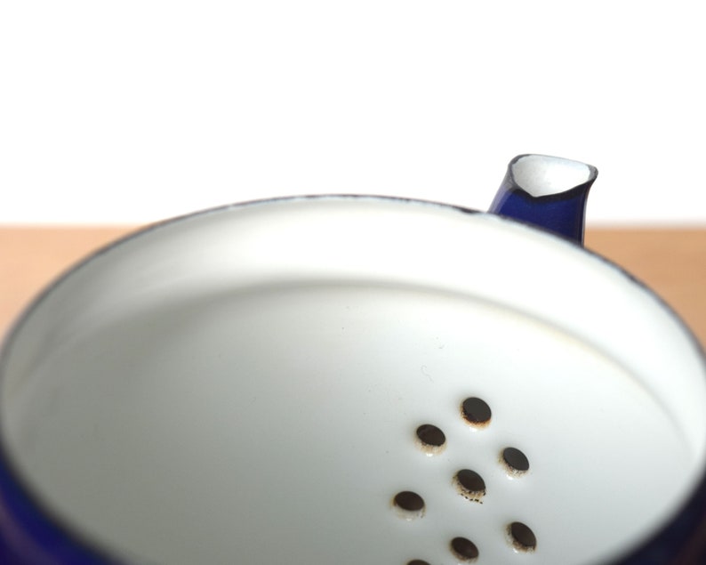 K&ouml;nnte beinhalten: Eine blaue Emailleteekanne mit wei&szlig;em Innenraum und einem Sieb mit mehreren L&ouml;chern. Die Teekanne ist aus Metall und hat ein klassisches Design.