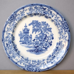 May include: A round, white ceramic plate with a blue floral and landscape design. The centre features a detailed blue illustration of a building and trees. The rim has a blue floral pattern. A classic piece for serving.