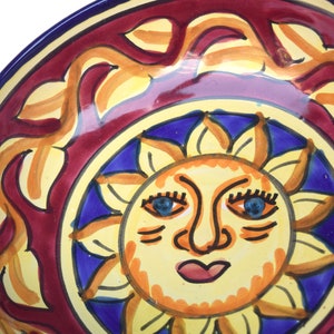 May include: A ceramic bowl featuring a red, yellow and blue sun design. The sun has a face with blue eyes, a yellow nose and a red mouth.