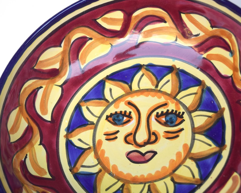 May include: A ceramic bowl featuring a red, yellow and blue sun design. The sun has a face with blue eyes, a yellow nose and a red mouth.