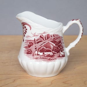 Może przedstawiać: Biały ceramiczny dzbanek z czerwonym wzorem transferowym. Dzbanek ma uchwyt i dzióbek. Wzór przedstawia krajobraz z napisem "Summer Morning". Dzbanek ma żłobioną podstawę i ozdobny uchwyt.