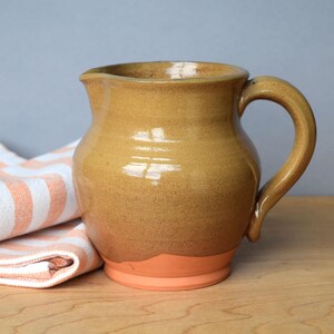 Pode incluir: Um jarro de cerâmica com um esmalte brilhante amarelo-mostarda na parte superior e uma base mate de cor terracota. O jarro apresenta um corpo arredondado, uma pega curva no lado direito e um pequeno bico vertedor no rebordo superior.
