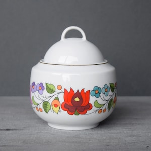 Könnte beinhalten: Weiße Zuckerdose aus Keramik mit Deckel, verziert mit einem farbenfrohen Blumendesign mit roten, orangefarbenen, gelben, grünen, blauen und violetten Blumen. Die Dose hat einen goldenen Rand am oberen Rand.