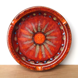 May include: A round, ceramic ashtray with a vibrant red and orange color scheme. The center features a sunburst design with orange, yellow, and green accents. The rim is decorated with blue and gold dots. The ashtray is approximately 6 inches in diameter.