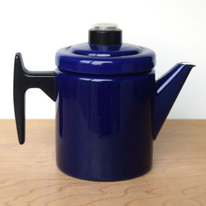 K&ouml;nnte beinhalten: Eine blaue Emaille-Kaffeekanne mit schwarzem Griff und Deckel. Die Kanne hat einen Ausguss an der Seite und einen kleinen Knopf auf dem Deckel.