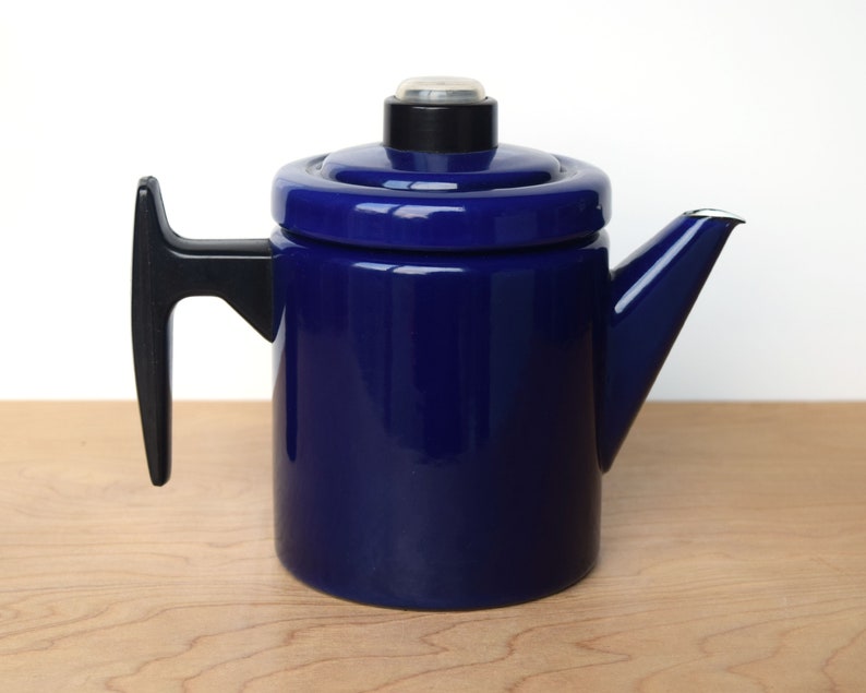 K&ouml;nnte beinhalten: Eine blaue Emaille-Kaffeekanne mit schwarzem Griff und Deckel. Die Kanne hat einen Ausguss an der Seite und einen kleinen Knopf auf dem Deckel.