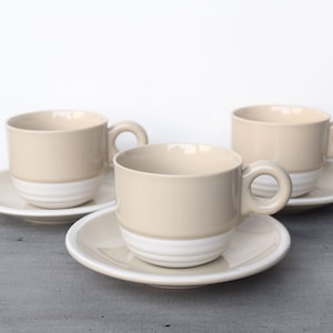 Op de afbeelding: Drie beige keramische theekopjes met witte banden rond de bodem, elk op een bijpassend schoteltje.