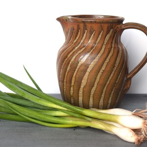 May include: A brown ceramic jug with a striped design sits on a grey surface next to a bunch of green spring onions.