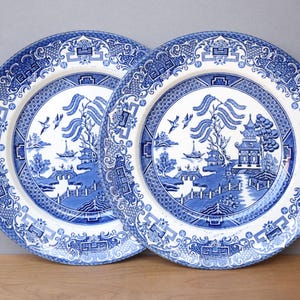 Puede incluir: Dos platos azules y blancos con el patrón Willow. Los platos presentan una escena detallada de paisaje con pagodas, puentes y pájaros. Los bordes tienen un borde decorativo. Los platos están sobre un fondo neutro.