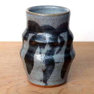 May include: A handcrafted ceramic vase featuring a grey and black abstract pattern. The vase has a cylindrical form with a gently widening rim and a textured finish. The dark, organic designs contrast with the light grey background.