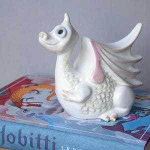 May include: A white ceramic dragon ornament with light blue eyes and pink wing details. The dragon sits on a book with the title "Hobitti". The figurine has a whimsical design, reminiscent of a fairytale creature.