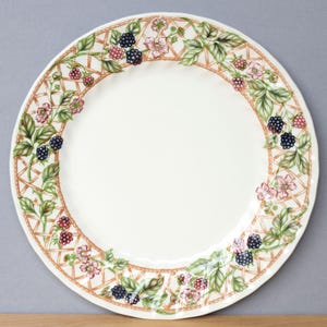 May include: A white ceramic plate with a decorative border featuring a brown lattice design, green leaves, pink flowers, and blackberries. The plate has a scalloped edge and is suitable for serving food.