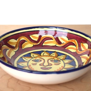 May include: A ceramic bowl featuring a sun design. The bowl is decorated with a yellow sun with a red face and blue eyes. The rim of the bowl is blue and white.