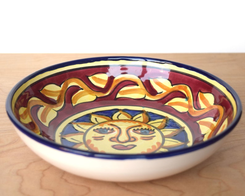May include: A ceramic bowl featuring a sun design. The bowl is decorated with a yellow sun with a red face and blue eyes. The rim of the bowl is blue and white.
