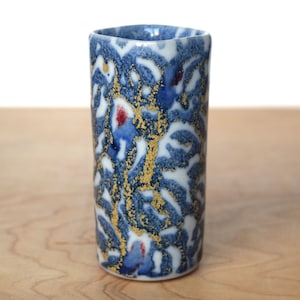 Può includere: Un vaso cilindrico in ceramica con un motivo astratto blu, bianco e oro. Il vaso ha una parte superiore leggermente svasata ed è posto su una superficie di legno chiaro. Il design include spruzzi di rosso.
