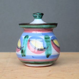 May include: A ceramic jar with a lid, featuring a colourful abstract design with green, blue, red, and yellow accents. The jar has a rounded shape and a smooth, glazed finish.