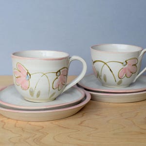 May include: Two ceramic teacups and saucers. The cups and saucers are white with pink floral designs and pink rims. The cups have handles. The saucers are round and have a pink rim. The background is a light blue.