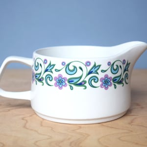 May include: White ceramic gravy boat with a blue and green floral pattern around the rim. The pattern includes blue swirls, green leaves, and purple flowers.