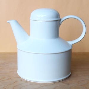 May include: A white ceramic teapot with a rounded body, a curved handle, and a lid. The spout extends from the top of the body. The teapot is sitting on a wooden surface.