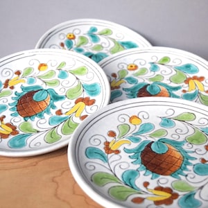 May include: Four white ceramic plates with a hand-painted floral design. The plates feature a brown centre, teal, green, and yellow leaves, and a black border. The plates are arranged on a wooden surface.