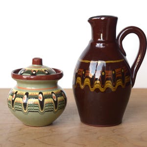 Pode incluir: Um jarro de cerâmica castanho com uma pega e um pequeno pote com tampa. Ambas as peças são decoradas com um padrão repetitivo de linhas onduladas amarelas, laranja e pretas sobre um fundo verde e castanho. O jarro tem um acabamento brilhante.