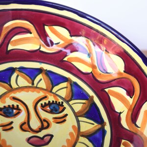May include: A ceramic bowl with a red, yellow and blue design. The bowl features a stylised sun with a face and a blue sky background. The rim of the bowl is blue.