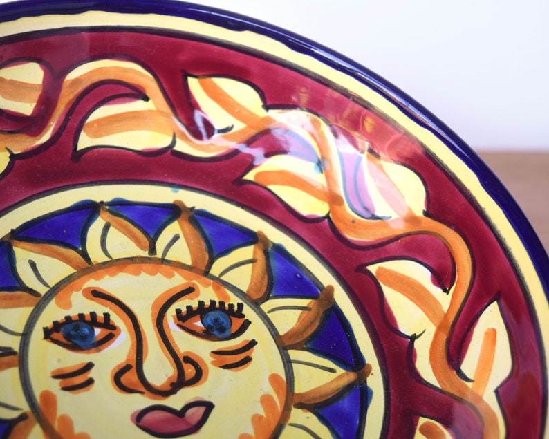 May include: A ceramic bowl with a red, yellow and blue design. The bowl features a stylised sun with a face and a blue sky background. The rim of the bowl is blue.