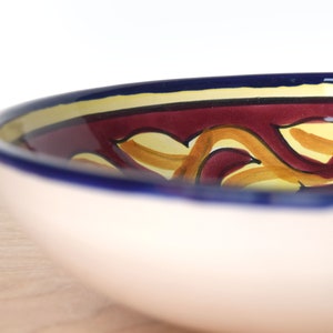 May include: A large, white ceramic bowl with a blue rim and a colourful design. The design features yellow and orange swirls on a burgundy background.