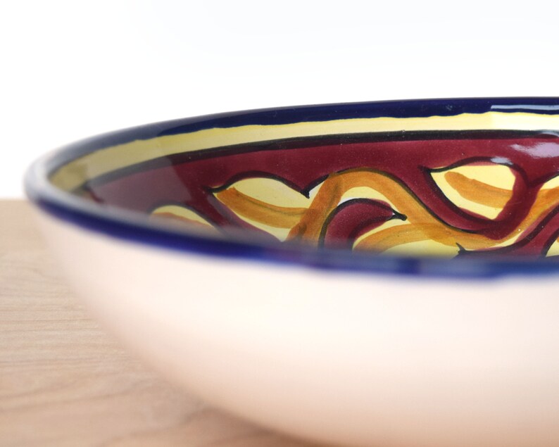 May include: A large, white ceramic bowl with a blue rim and a colourful design. The design features yellow and orange swirls on a burgundy background.