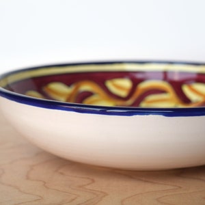 May include: A white ceramic bowl with a blue rim and a colourful design of yellow and red swirls. The bowl is on a wooden surface.
