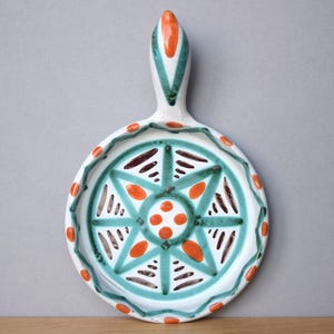 Könnte beinhalten: Weißes Keramik-Serviertablett mit Griff, mit einem türkisfarbenen und orangefarbenen Sternmuster in der Mitte. Der Rand ist mit orangefarbenen Punkten und grünen Akzenten verziert. Das Tablett hat einen Durchmesser von ca. 25 cm.