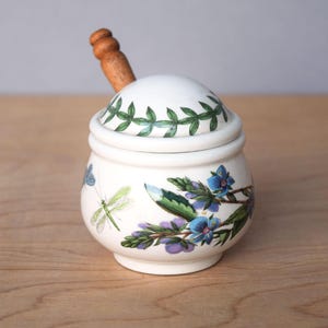 May include: A white ceramic sugar bowl with a lid and a wooden spoon. The bowl is decorated with green leaves, blue flowers, and dragonfly illustrations. The spoon is inserted into the lid.