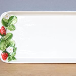 May include: A rectangular white ceramic serving platter with rounded corners. The left side is decorated with a strawberry motif, featuring red strawberries, green leaves, and white flowers. The platter is designed for serving food.