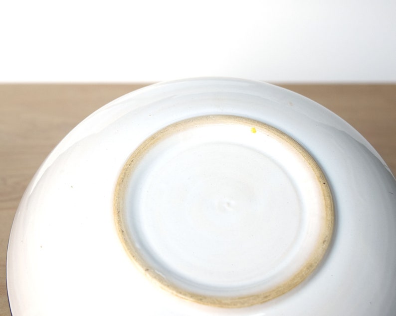 May include: White ceramic bowl with a slightly raised rim and a brown glazed base. The bowl is empty and sitting on a wooden surface.