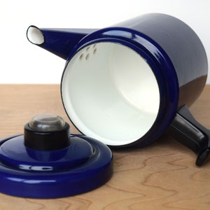 K&ouml;nnte beinhalten: Eine blaue Emaille-Kaffeekanne mit schwarzem Griff und wei&szlig;em Innenraum. Der Deckel ist abgenommen und die Kanne steht auf einer Holzoberfl&auml;che.
