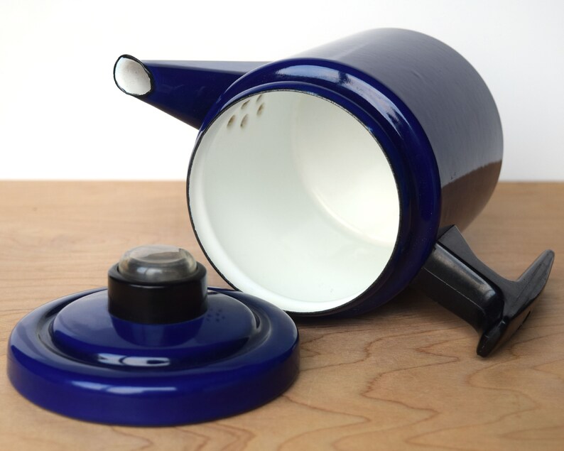 K&ouml;nnte beinhalten: Eine blaue Emaille-Kaffeekanne mit schwarzem Griff und wei&szlig;em Innenraum. Der Deckel ist abgenommen und die Kanne steht auf einer Holzoberfl&auml;che.