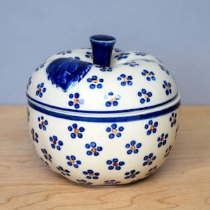 Puede incluir: Un tarro de cerámica con forma de manzana con tapa. El tarro es blanco con patrones florales azules y un borde azul. La tapa tiene un tallo azul y un diseño de hoja azul. Colocado sobre una superficie de madera.