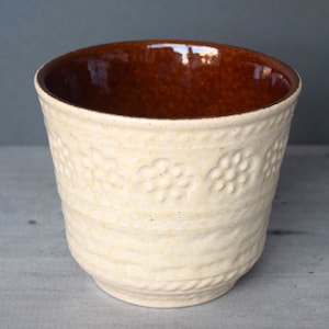 Op de afbeelding: Een witte keramische pot met een bruin geglazuurd interieur. De pot heeft een textuurontwerp met reliëfbloemen aan de buitenkant.