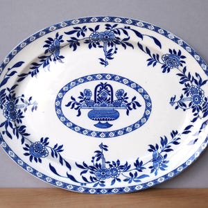 May include: Oval white serving platter with a blue floral design. The platter features a central oval with a basket of flowers and a border of flowers and leaves. The rim has a blue geometric pattern.