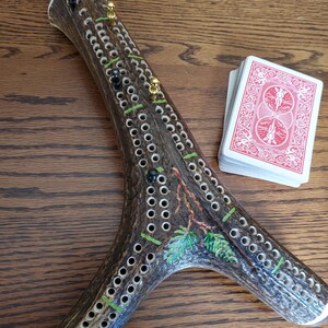 May include: A Y-shaped cribbage board crafted from a textured brown antler, featuring rows of drilled holes for scoring, small gold and black metal pegs, and a painted green pine branch design, accompanied by a deck of red and white playing cards.