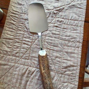 May include: A stainless steel pie server with a natural antler handle. The server is resting on a quilted placemat with a repeating pattern of small red squares on a beige background. The handle is a dark brown color.